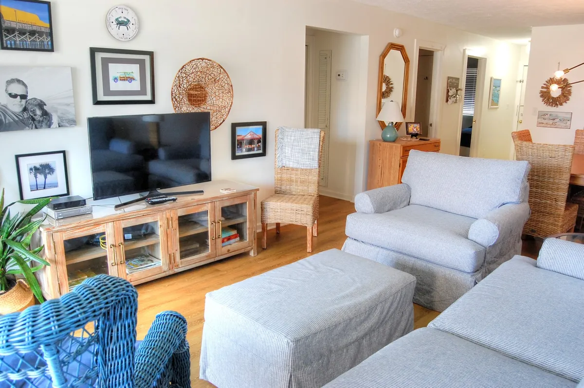 A cozy living room scene with a comfortable seating arrangement, a television, decorative wall art, and woven chairs. The mood is relaxed and inviting, suggesting movie nights, board games, or quiet moments with friends and family.