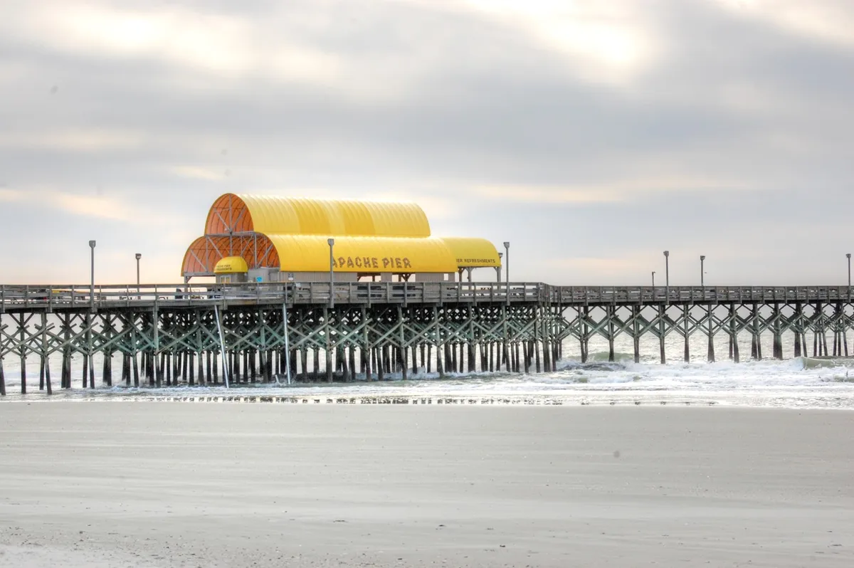 A sweeping view of Apache Pier stretching over the ocean waves, featuring its distinctive yellow roof and wooden planks, with the gentle motion of water beneath and the open sky above. The scene exudes tranquility and hints at sunset or early evening light, inviting visitors to stroll or fish.