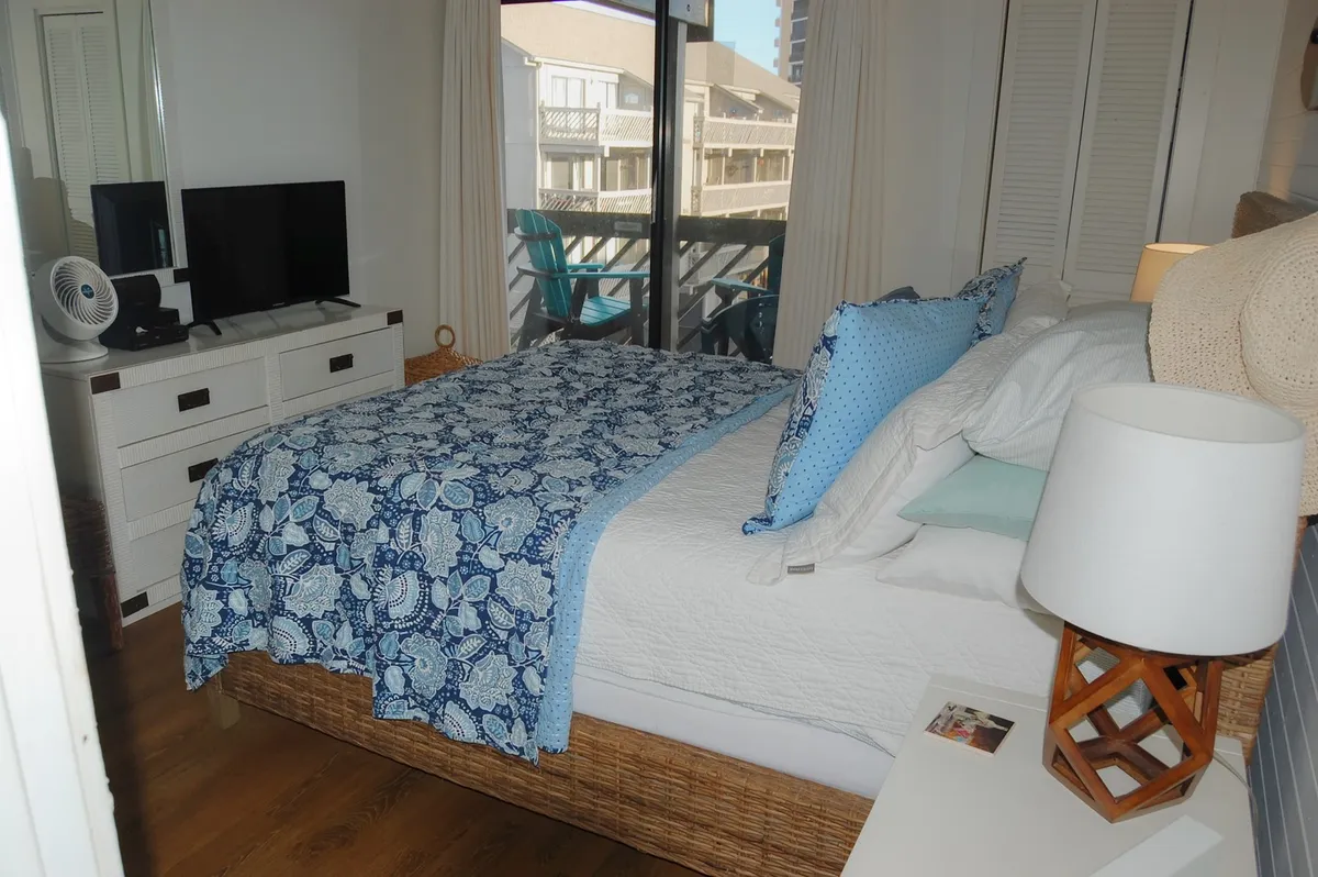 This image features a bedroom with a bed dressed in a blue patterned blanket, a dresser with a TV, and large windows offering a view of a balcony. The room is bathed in natural light, conveying a serene and inviting atmosphere ideal for rest and relaxation. Details like the fresh linens, cozy textiles, and ample storage on the dresser directly support the themes of comfort and thoughtful amenities.