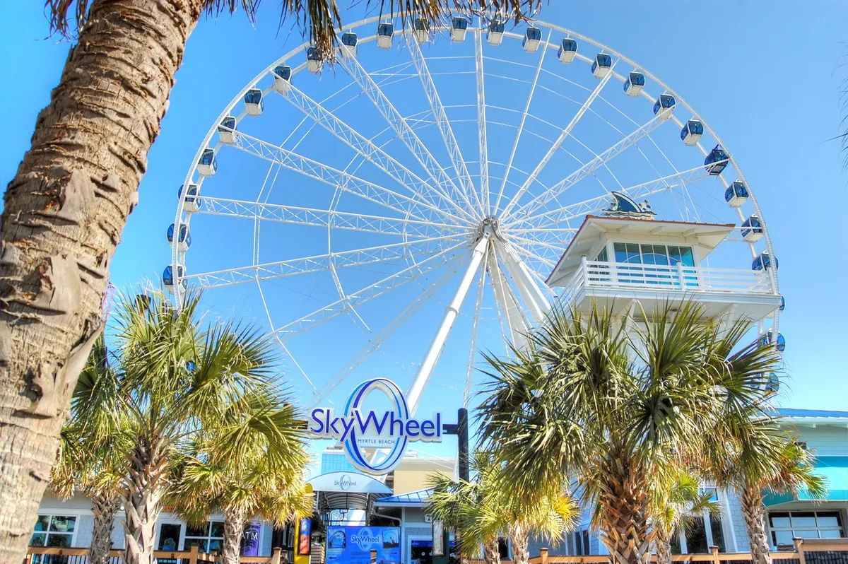 This image features the iconic Myrtle Beach SkyWheel, palm trees, and the entrance building. The scene is lively and bright, with people preparing to ride the towering ferris wheel, which is a well-known local attraction included in the Free Fun Program.