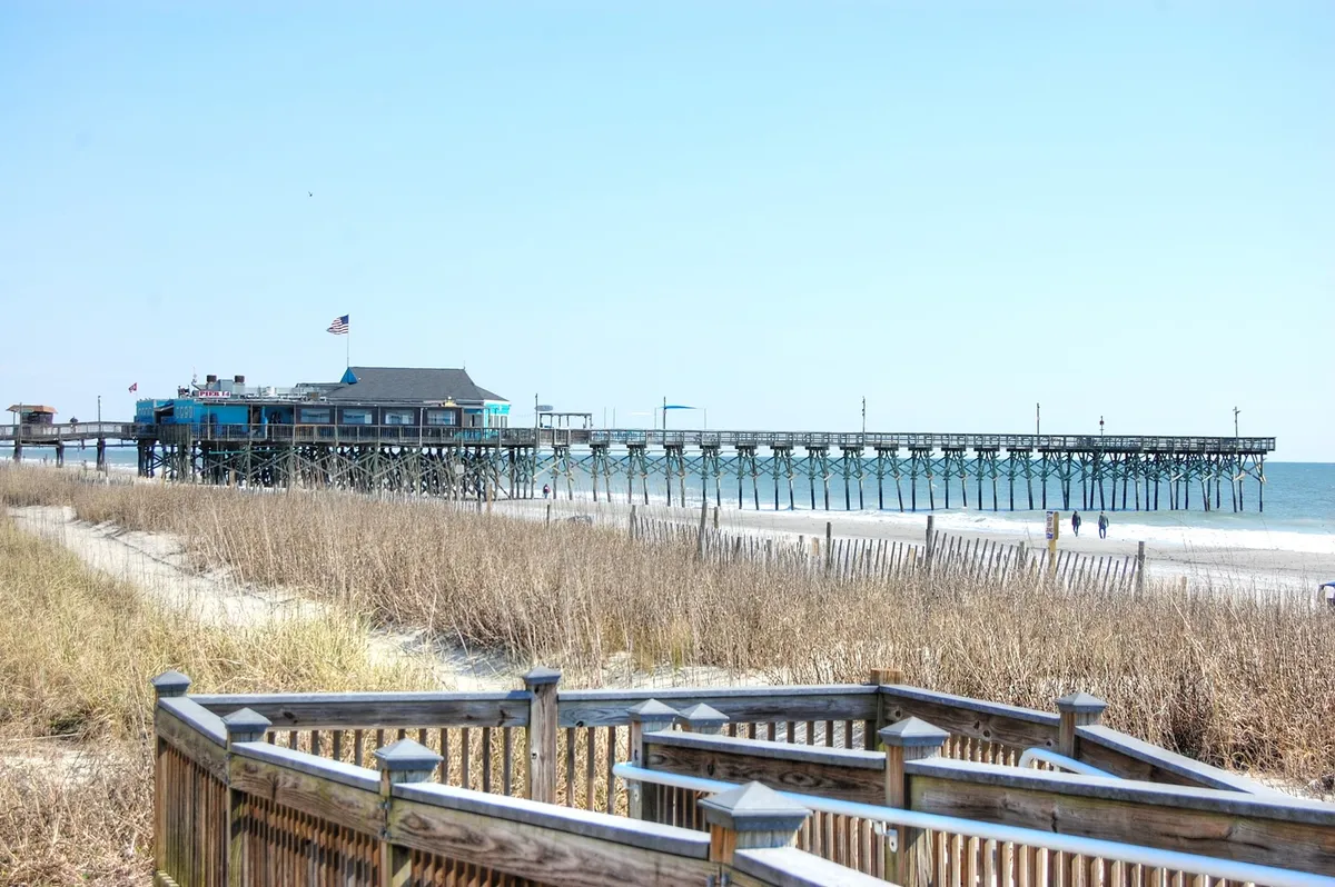 A scenic view of a wooden pier stretching out over the ocean, with sand dunes and the expansive sky in the background. The beach appears clean and inviting, showcasing both the sandy shoreline and the lure of the ocean just steps away.
