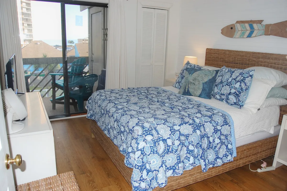 A serene bedroom with a blue patterned quilt on the bed and a sliding glass door leading out to a private balcony. The balcony is partially visible, hinting at opportunities for morning coffee or evening conversations while enjoying fresh coastal breezes.