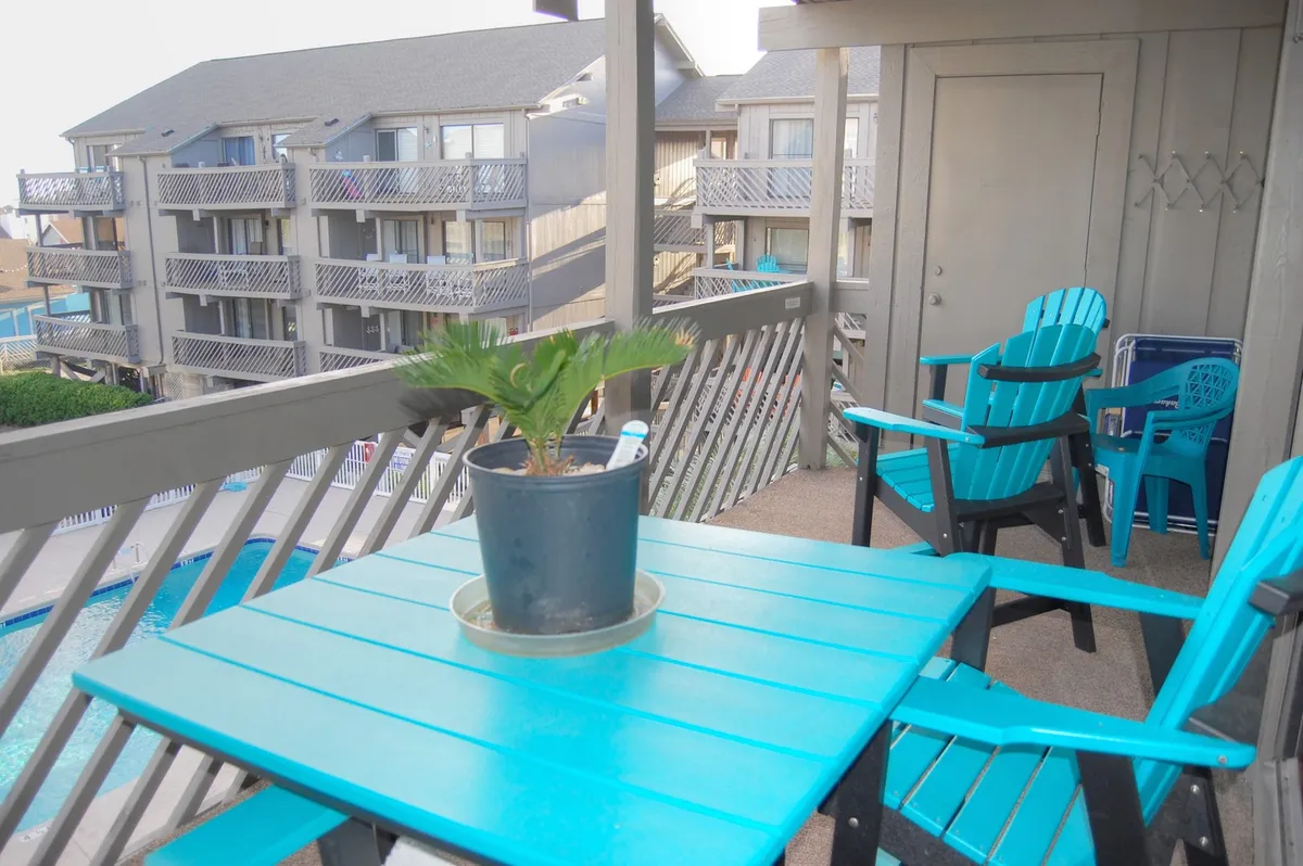 An exterior shot featuring a turquoise table and chairs set on a sun deck, with a swimming pool visible in the background. The potted plant and bright setting suggest a perfect spot for morning coffee, reading, or socializing with friends as the sun rises or sets.