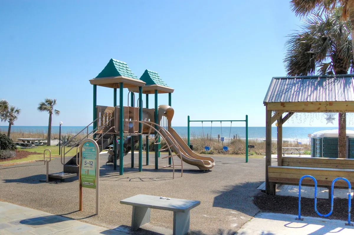 A family-friendly playground set against a backdrop of palm trees and the beach, with swings and play equipment visible. The ocean can be seen just beyond the playground, creating a joyful, active, and scenic environment.