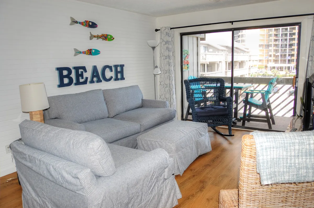 The image showcases a bright, inviting living area with a comfy sofa, playful decorative fish accents, a cheerful 'BEACH' sign, and a balcony offering a glimpse of sunrise views. A stylish blue chair adds a pop of color, suggesting a perfect spot to unwind or enjoy a book. The overall ambiance feels cheerful and family-friendly, ideal for relaxation and social gatherings.