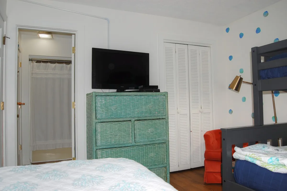 This image offers a broader view of the second bedroom, featuring a blue bunk bed, green wicker dresser for storage, an orange chair, and a TV. The vibrant colors and casual furniture invite relaxation, reading, or movie nights, making it a versatile space that suits families or groups who want to spread out.