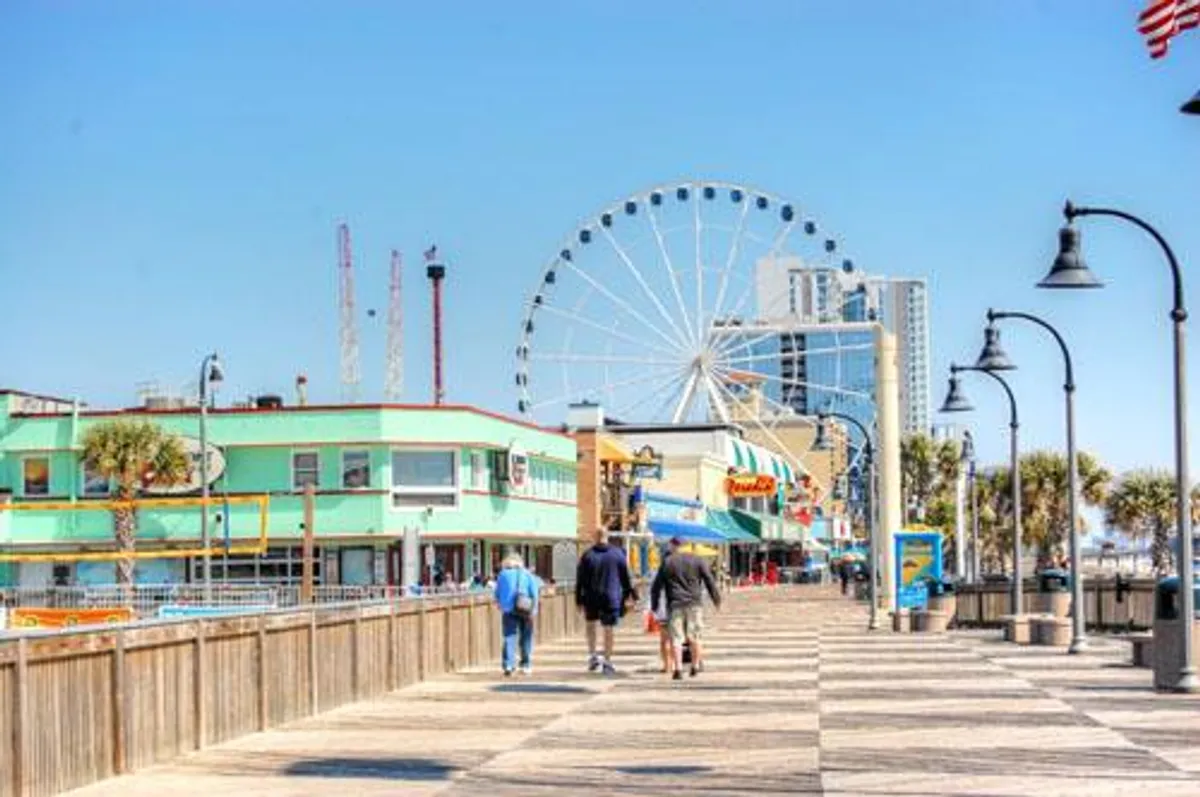 A bright exterior view of a bustling boardwalk with a colorful ferris wheel, shops, and groups of people enjoying the lively scene. The festive mood and iconic attractions make it an inviting representation of Myrtle Beach's entertainment options.