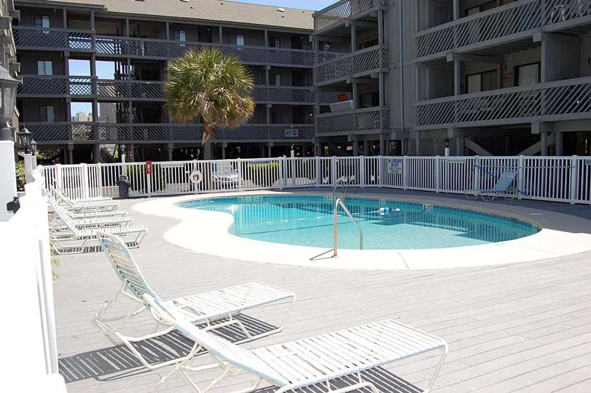 A sparkling outdoor pool surrounded by palm trees, lounge chairs, and a secure fence, basking in inviting sunlight. The pool area hints at relaxation and leisure opportunities available even during winter months.