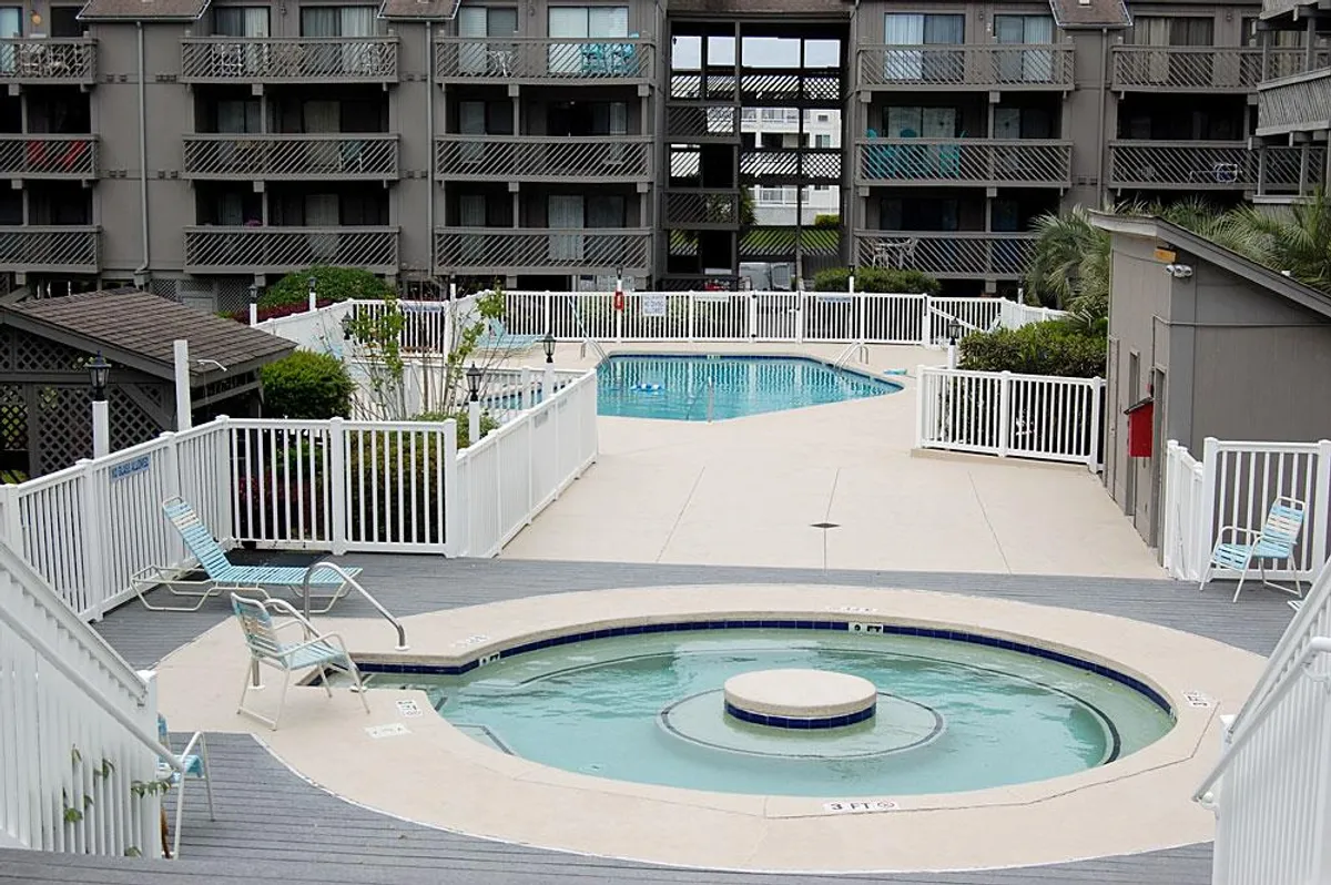 An inviting outdoor swimming pool area with a hot tub and several lounge chairs arranged around the water. The setting is sunny and bright, with a clear view of the pool and places to relax or socialize.