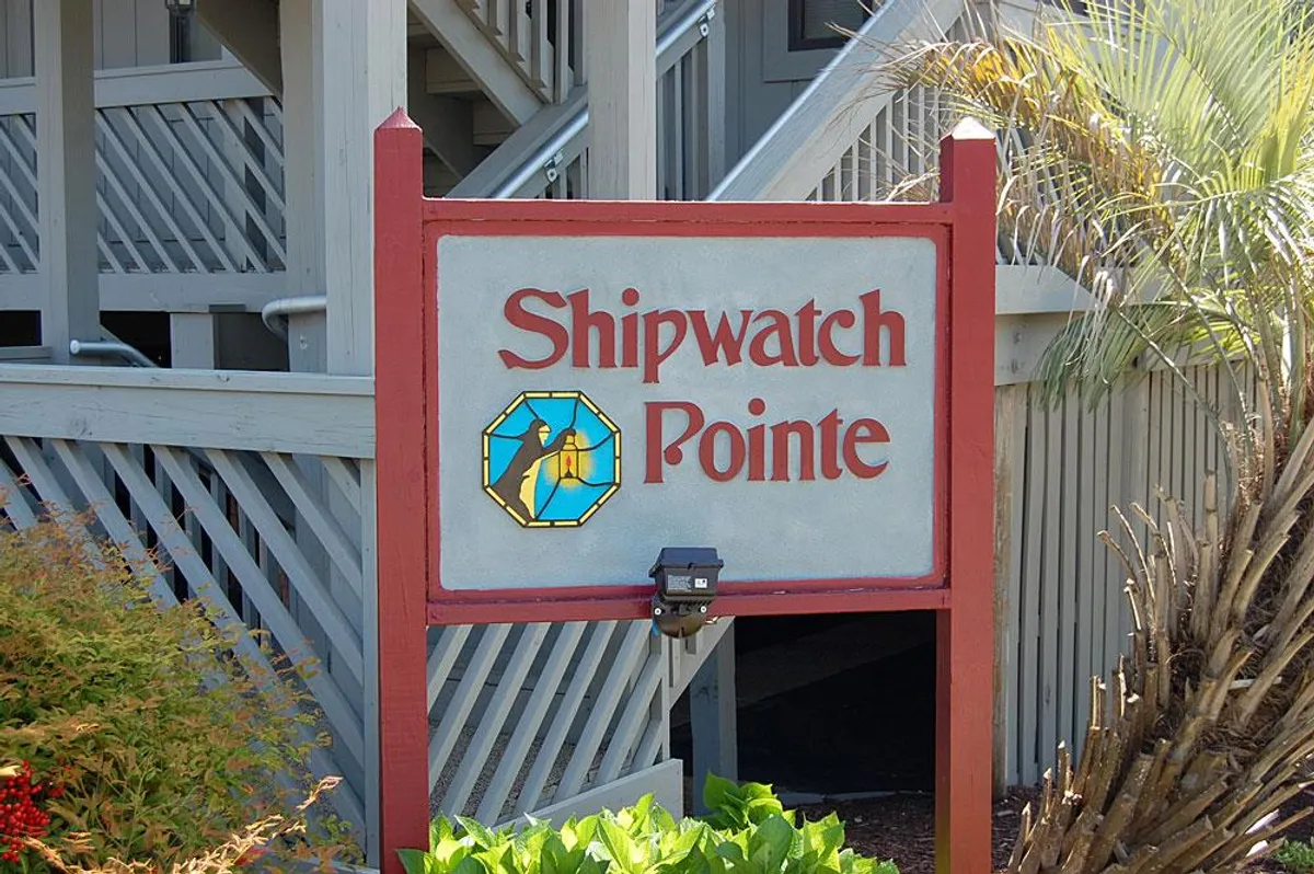 This exterior image features the welcoming signage of Shipwatch Pointe, surrounded by lush landscaping and a staircase leading to the upper levels. The scene is bright and friendly, immediately signaling arrival at a comfortable seasonal getaway.