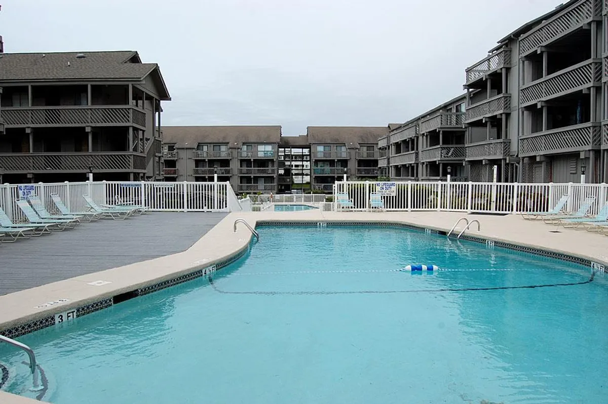 A lively exterior shot featuring a sparkling swimming pool surrounded by lounge chairs and the condo buildings. The space is set up for socializing, sunbathing, and group fun, with plenty of room for both relaxation and playful activities.