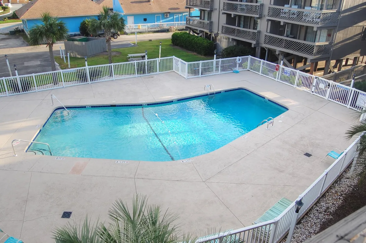 A lively exterior scene with a large swimming pool surrounded by palm trees, a fenced-in area, and adjacent condo buildings. Lounge chairs are scattered around the pool, inviting guests to relax and socialize in the sun. The space feels open and designed for community enjoyment.